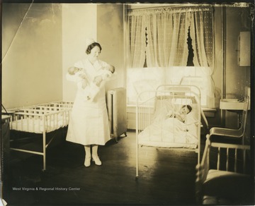 A nurse holds a baby in each arm. On the right of the photograph, a child sleeps in a crib.