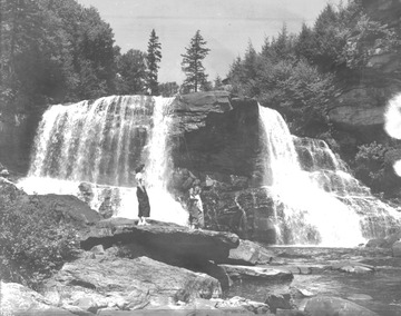 A woman stands on a rock as she observe the scenery. In the background is a waterfall. 