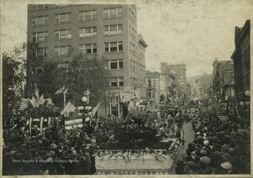 A crowd watches from the street as parade floats make their way down High Street. On the right, in front of the courthouse, is likely the grandstand.