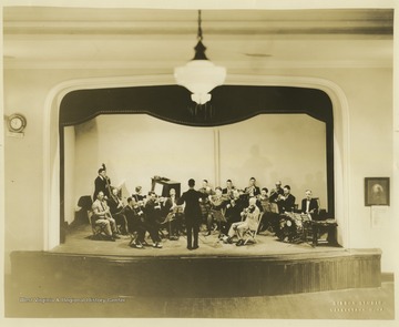 The man standing in the middle of the stage directs the musical group which consists of brass, string, woodwind, and percussion musicians inside the Women's Christian Temperance Union Community Building.