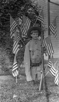 Son of photographer James Edwin Green, 9-year-old James Edwin Green, Jr. (b. 1904) poses in a Spanish War uniform while holding a rifle. He is surrounded by American flags, celebrating the Fourth of July.