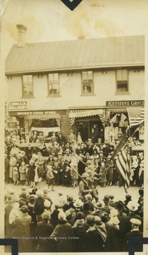 A group of soldiers march in the parade down High Street.  A cigar store and a grocery are visible in the background.