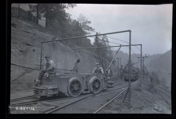 Four men stand outside of mine with loaded mine car ready to be hitched to electric engine car in front.  