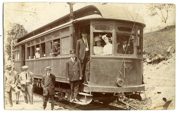 A conductor poses on the steps of a street car filled with passengers.
