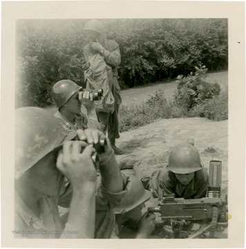Men look down-range with binoculars as another man loads machine gun.