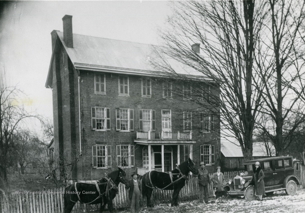 Two men standing in front of the home, each with a horse, and two women standing in front of a car with a pet dog.