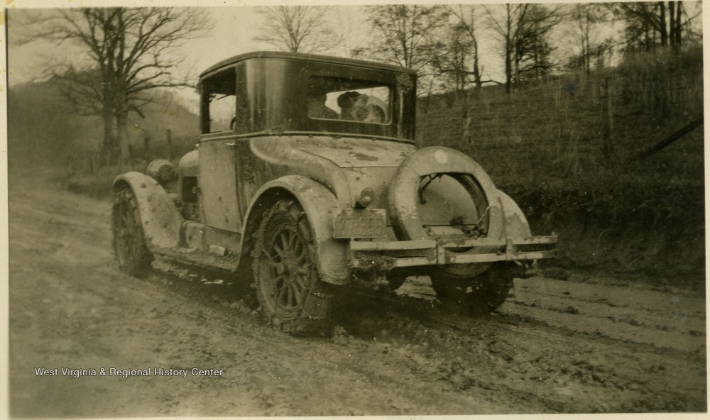 A vehicle with chains on the tires to navigate an unpaved muddy road. It has a West Virginia license plate and is thought to have belonged to the Mathers/Barrick family.