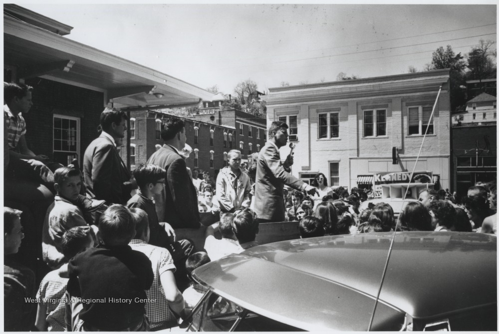 Kennedy speaks to a crowd gathered around the podium. To Kennedy's right is brother, Ted (Edward) Kennedy, and West Virginia political figure, A. James Manchin.