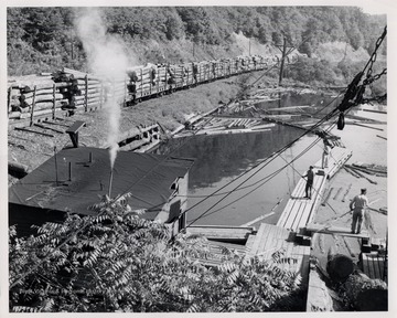 Men direct floating logs on the pond as a log train passes.
