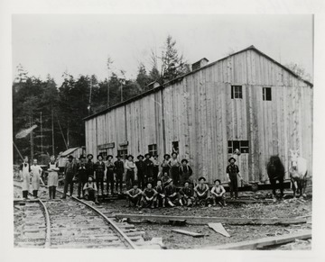 Large group of men in front of a building beside a railroad track.  
