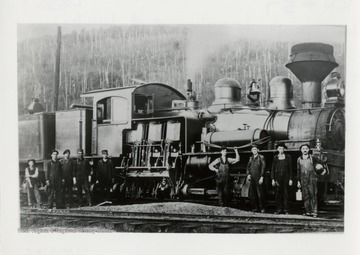 Side view of train engine with nine crew members standing in front of it.  