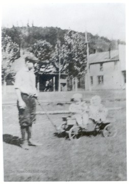 Small boy pulling a wagon with two kids through the grass.