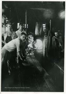 Four glass workers in factory standing near the furnace. One worker holding pipe and glass cylinder out of furnace.