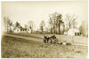 Man plowing a field with a tractor; house, car, and barn in background. 