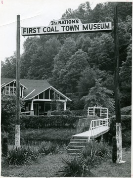 The 'First Coal Town Museum' seated at the bottom a hill.  A small bridge spans a rock ledge adjacent the entrance of the museum.