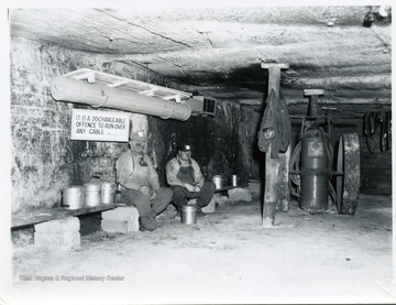 Two men sitting on a bench in a mine in an underground mine.  Sign reads, 'It is a dischargeable offence to run over any cable.'