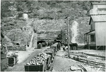 Coal carts line up in front of the mine entrance full of coal.  Men and horses stand beside them.