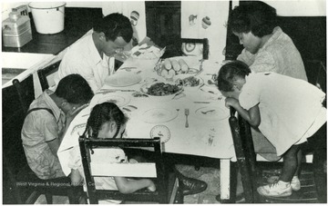 Family saying prayer at the dinner table. From Book: A Medical Survey of the Bituminous Coal Industry, a report of the coal mine administration. 'Washington: 1947.'