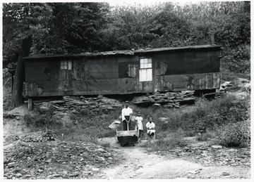 African-American miner holding a wooden cart and two children stand below their home.
