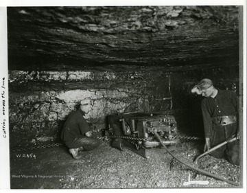 Two miners using a machine to cut across the face of a wall in a mine.