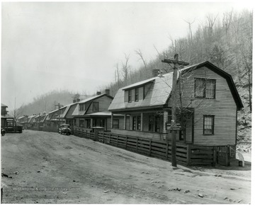 Row of houses along a dirt road with a few cars parked out front. Sign reads: 'Notice, speed limit 15 miles per hour, persons exceeding this limit will be prosecuted'.