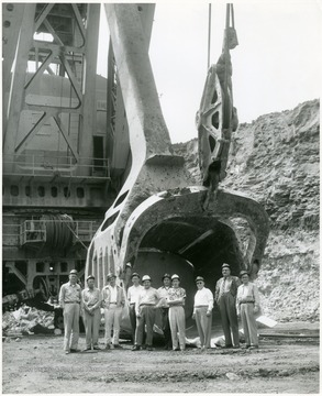 Men in hard hats stand in front of giant shovel scoop.