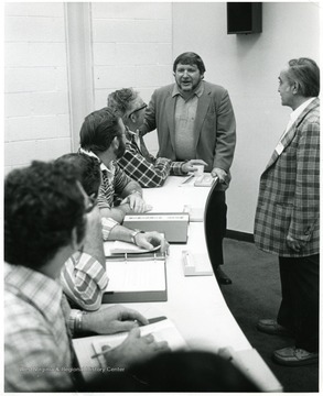 A group of men sit and stand around a table.