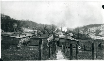 Wooden walkway leading to a group of barracks. Some snow on hillside in distance.