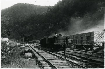 One man is sitting on a filled coal car in front of the coke ovens while another is standing on the tracks.