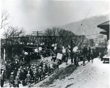 Miners crowd the streets and cross a metal bridge in a large parade. Many hold U. M. W. of A. signs.