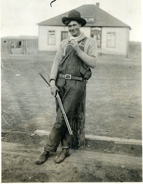 "Thug" leaning against a tree trunk holding a gun, saloon in background.