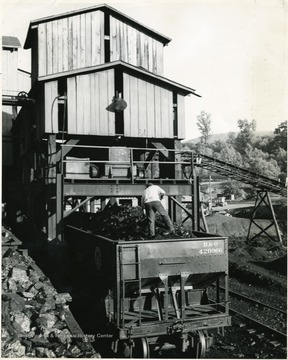 A man stands atop a filled coal car. 'Loading coal into railroad gondolas is made easy by structures called 'tipples.' The pictured tipple is typical of those to be found throughout the rich bituminous coal fields of southern West Virginia.'