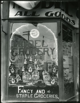 Grocery store window with advertisements for 'fancy and staple groceries' sold inside.