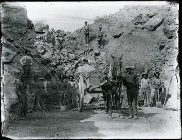 African-American miners with picks and shovels and a horse drawn cart stand in front of a large pile of boulders.