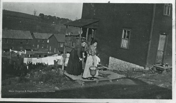 Two women pumping water into a bucket beside a house.