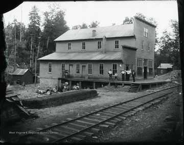 Men standing on porch of a building next to railroad tracks. There are empty barrels underneath the porch.