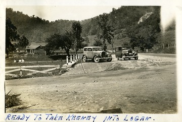Two cars parked alongside a road.  Cabin visible in left background.  Photograph from Joe Ozanic scrapbook.
