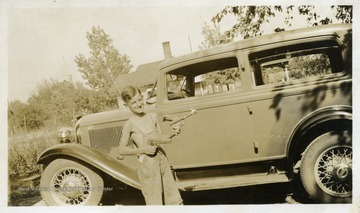 Small boy wearing overalls, standing with toy guns, in front of a car. 