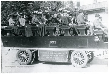 Group of people sitting on benches on a wagon. Sign on the side says 'All Around Washington Auto Co., Office 14th [and] PA. Ave. Phone 2261.'