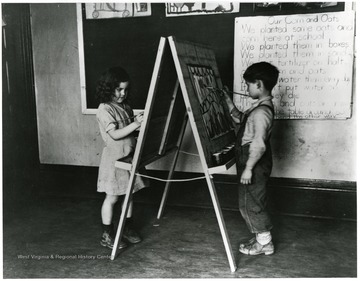 'First grade boy and girl at drawing board in school room at Reedsville.'