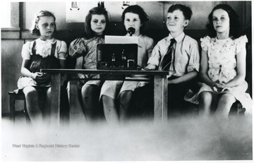 Children sitting behind a table.   Girl in center speaks into a microphone.