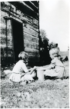 Two girls seated with a bucket in between them next to a log cabin at Arthurdale.