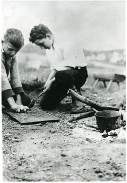 Two boys crouched down to the ground.  One is holding a ladle over the other's hand and a rectangular shaped piece of wood.  A kettle sits on the side of a fire.