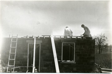 Two men work atop a brick building installing a roof.