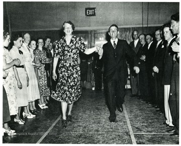 Eleanor Roosevelt and man move happily down collumn of people in large auditorium in Arthurdale W. Va. From The Eleanor Roosevelt We Remember page 78. 'A print from the FDR Library Collection. This print is furnished for your file and must not be reproduced without the owner's permission. Owner: Wide World.'