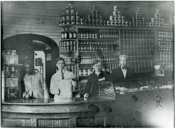 Three young men and an African-American man lean against the counter in front of shelves lines with canned and bottled goods.