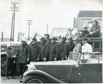 Eleven men and one women standing in front of a fire truck.  Three men are sitting in the fire truck.  