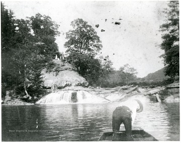 Man stands in a boat near where logs are caught in some rapids.