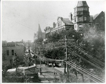 Two men are crossing a dirt street in Charleston, West Virginia in the early 1900s. On one side of the street are businesses while on the other side of the street are houses. Above the street are lots of telephone wires.