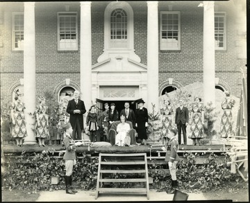 On stage in front of the school is a girl with a crown with other people around her on both sides.  